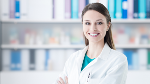 A healthcare professional is smiling at the camera with her arms crossed. She is standing in front of a bookshelf in a clinical setting.