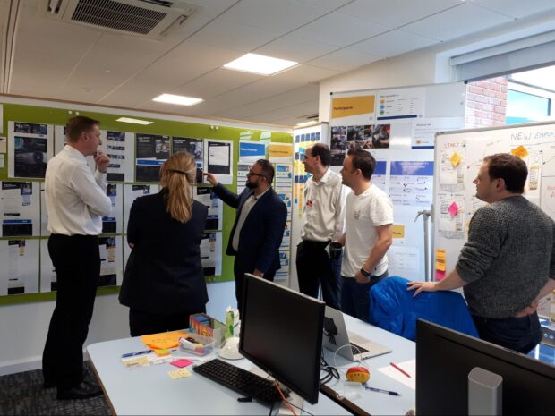 Six people standing aroud a wall pointing at various different posters and screenshots.