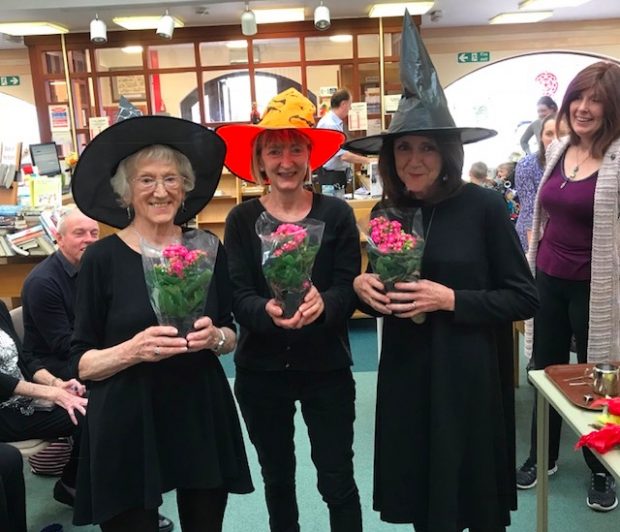 Photo of 3 women in a library - dressed in black and wearing pointed witches hats