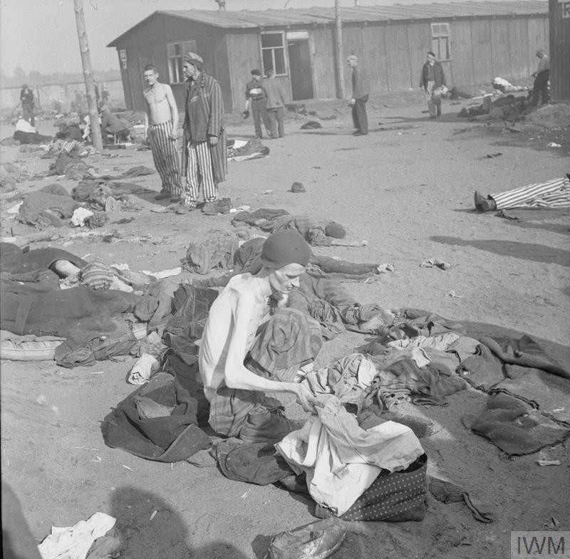 An emaciated prisoner of Bergen Belsen concentration camp delouses a  pile of striped clothing, there are prone bodies lying on the floor around him. 