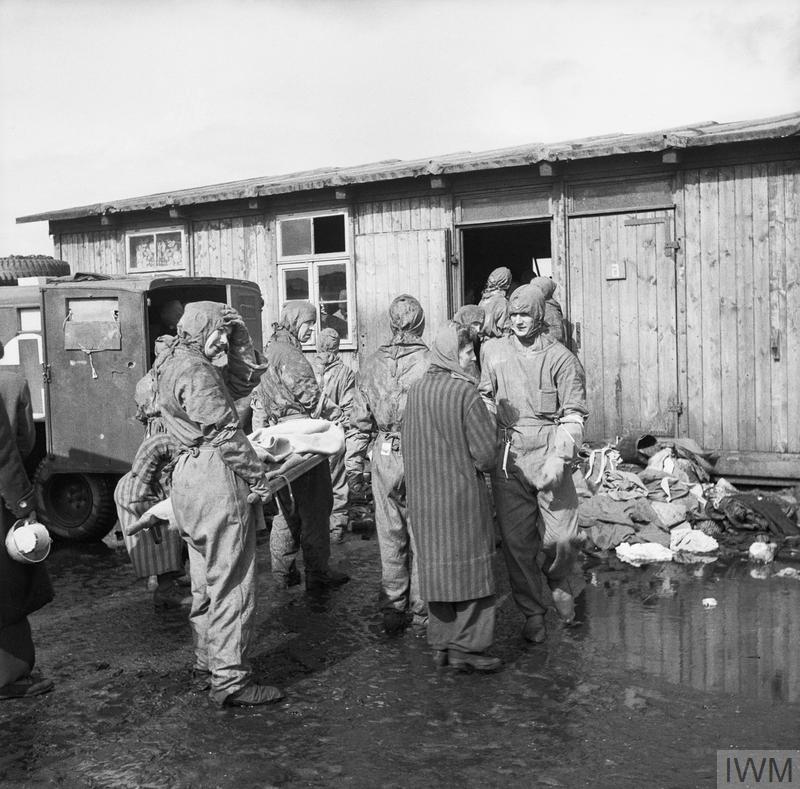 British soldiers wearing protective overalls and head coverings, line up in front of a wooden hut at Bergen Belsen, some carry a stretcher to evacuate the sick.
