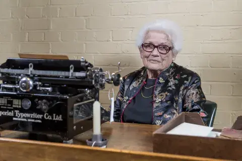 Joy Hunter, a typist in the Churchill War Rooms during the Second World War, sits behind a desk 