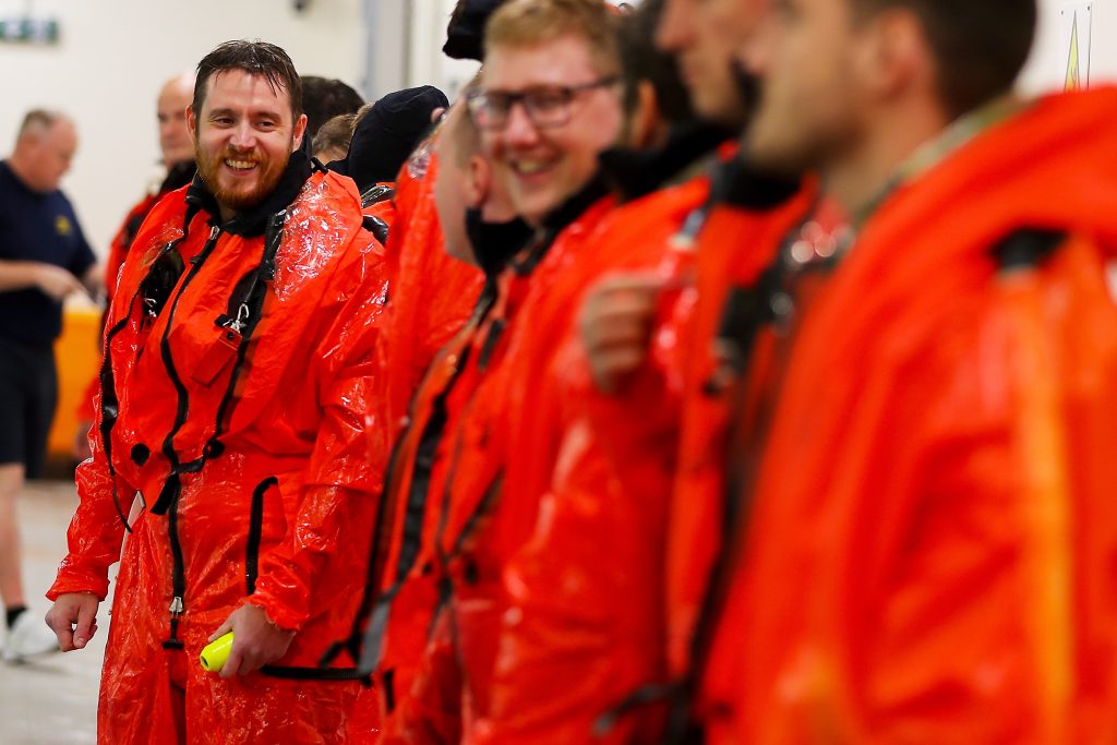 A line of sailors in bright orange dry suits. Only one is in focus, smiling at a companion next to him. His dry suit is wet. 