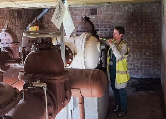 Charlie is wearing a yellow sleeveless vis-me jacket and is taking a gas meter reading with a small device. She is pictured next to a big white pipe and a brown one.