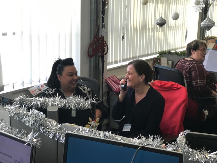 Wendy sits at a desk on the phone. She is smiling at a woman sitting next to her. Behind them is another woman, also sat at a computer. The office has tinsel and Christmas decorations.