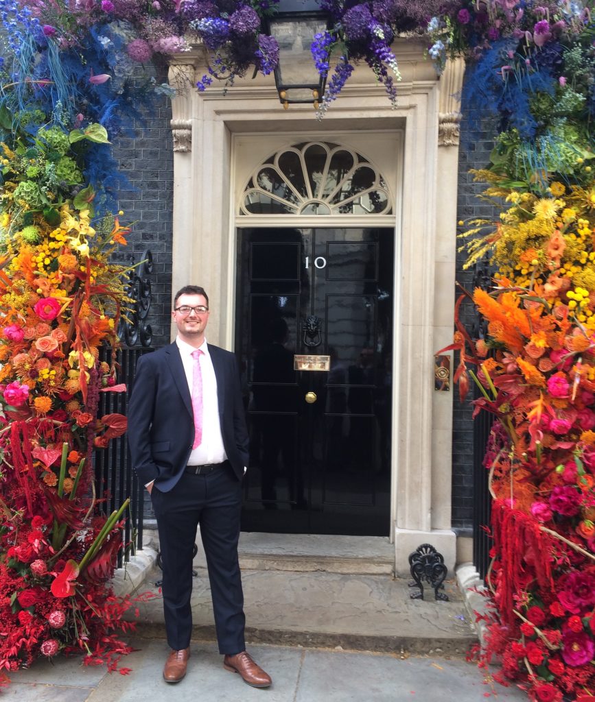 Nathan is standing outside of Number 10 Downing Street which has flowers all around the doorway. The flowers are in the pride colours of red, yellow, green, blue, purple. Nathan is wearing a black suit with a white shirt and pink tie and is in smart brown shoes.