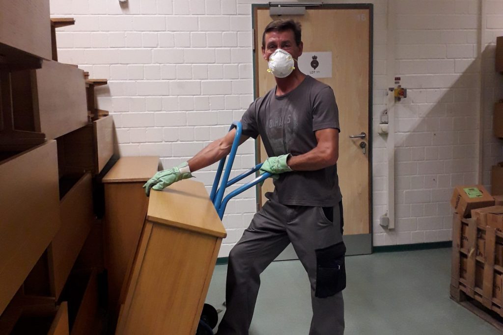 A man wearing gloves and a white face mask is moving a piece of furniture onto a blue trolley.