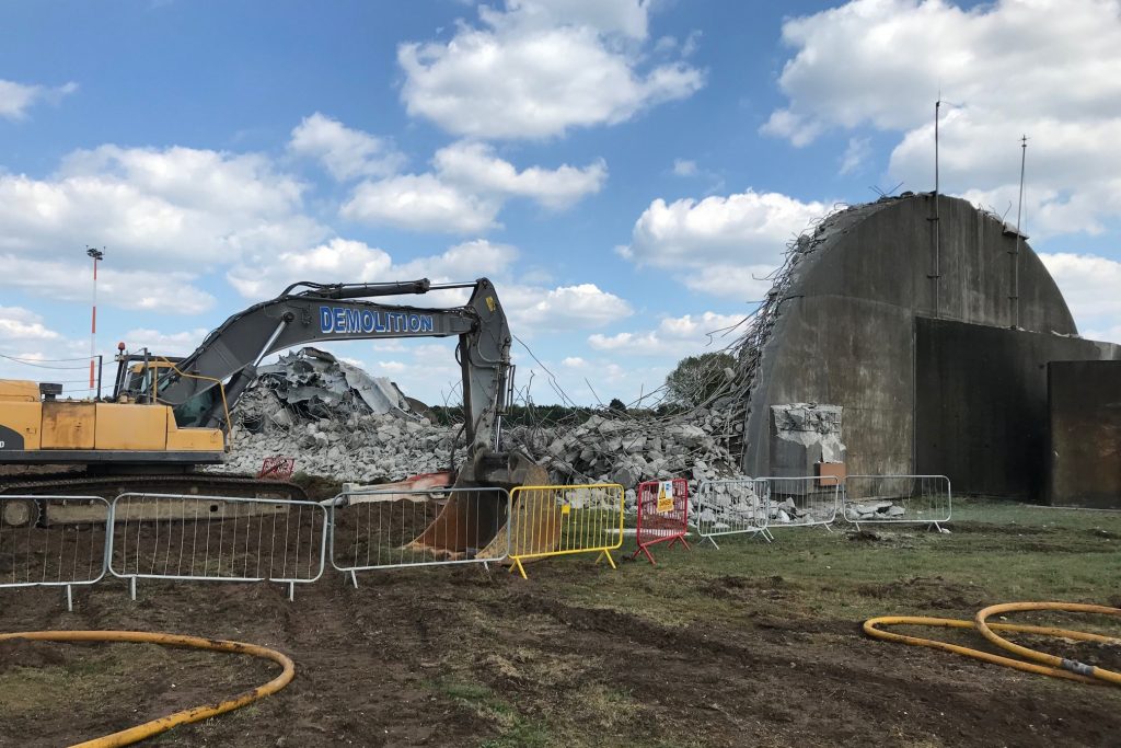 A large digger behind a fence, with a pile of rubble and a remaining semi-circular concrete wall which still stands.