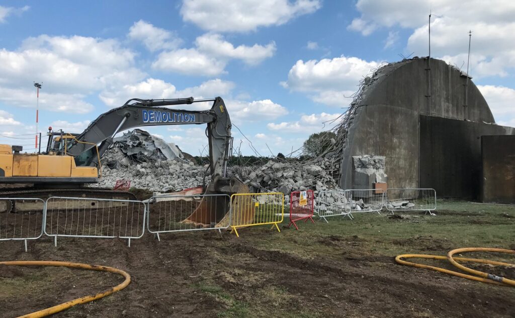 A demolition vehicle is located on the left with removal of construction materials beside it. A building is located on the left that is being demolished.