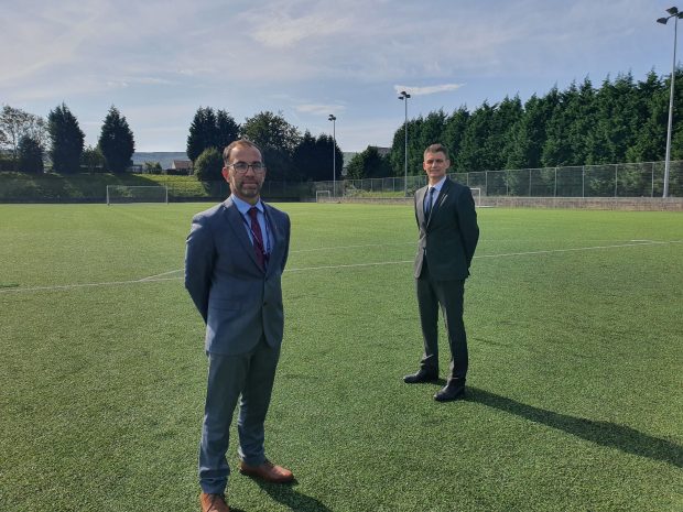 Two men stand 2m apart on a school field facing the camera