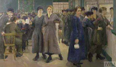 Crowded canteen of female workers. A counter and tables are occupied with workers in overalls and cloth caps.