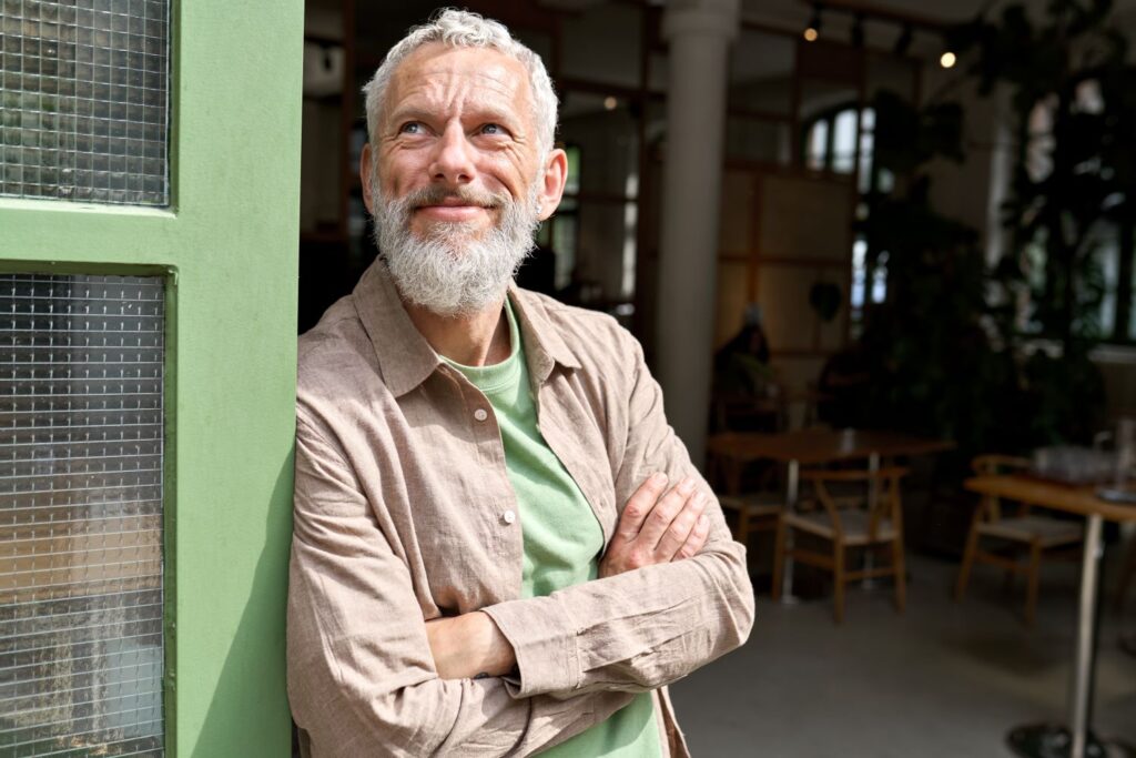 Smiling man with a beard and white hair stands in a doorway with his arms folded