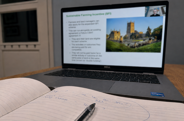 A Dell laptop on a wooden table displays an online presentation titled “Sustainable Farming Incentive (SFI)” with bullet points on the left and a photo of sheep grazing near a church on the right. In front of the laptop, an open lined notebook with handwritten notes and a black pen rests on the table, suggesting someone is taking notes during a webinar.