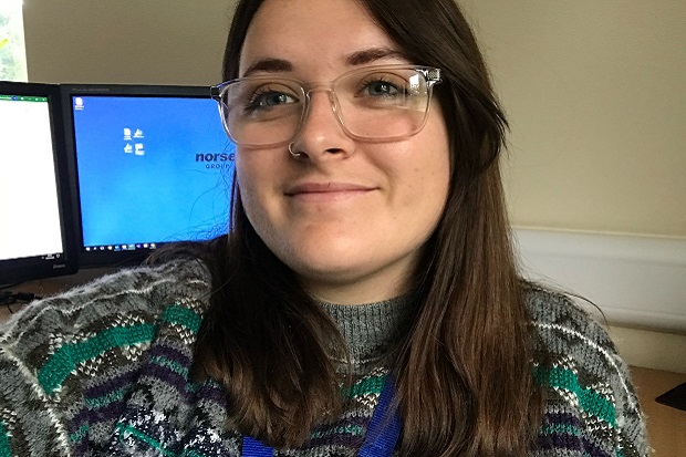A girl with glasses, long dark hair, and wearing a patterned jumper, with a computer screen behind her.