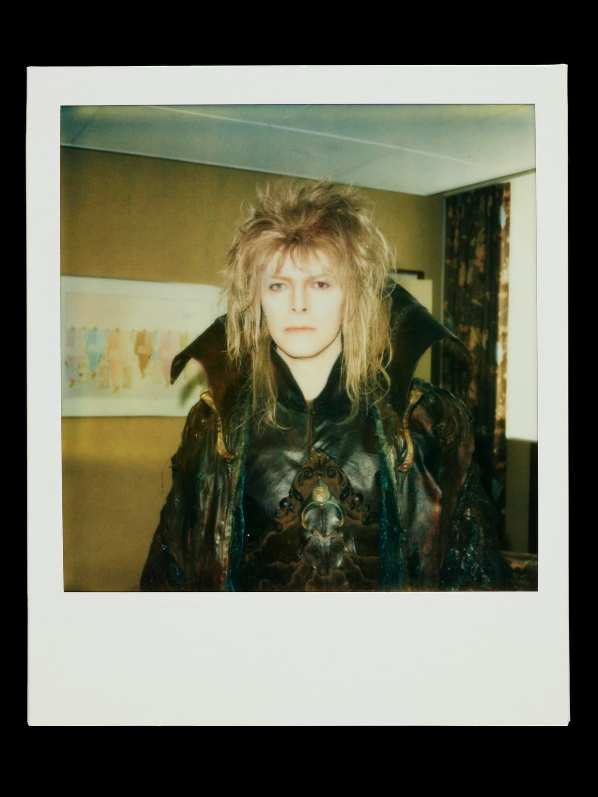 David Bowie with spiky hair wearing ornate, medieval-style costume with high collar, standing in a room with a painting and floral curtains.