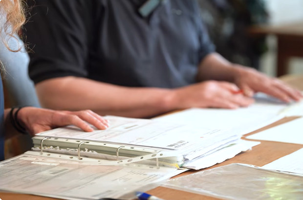 Close-up of two people at a desk reviewing documents, with hands visible over a ring binder folder containing printed papers, suggesting paperwork or application forms being checked together.