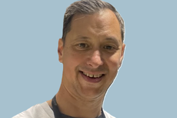A man with short swept back hair, against a blue background.