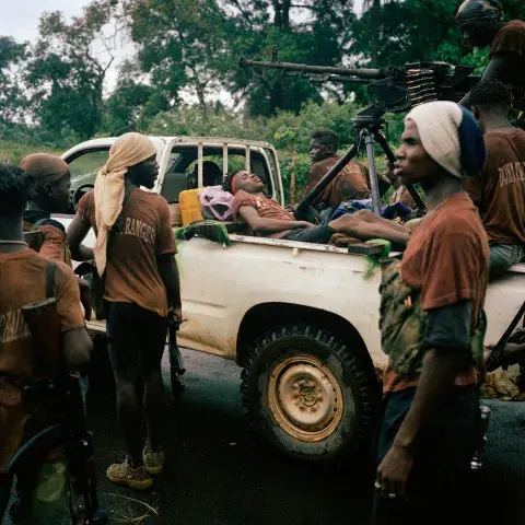 A group of rebel fighters in and around a truck. A fighter, wounded in an ambush, lays in the back of a pick up truck beside a mounted machine gun.