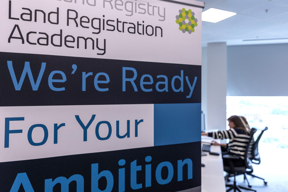 A banner saying 'Land Registration Academy' and 'We're ready for your ambition', with caseworkers glimpsed at a desk behind.