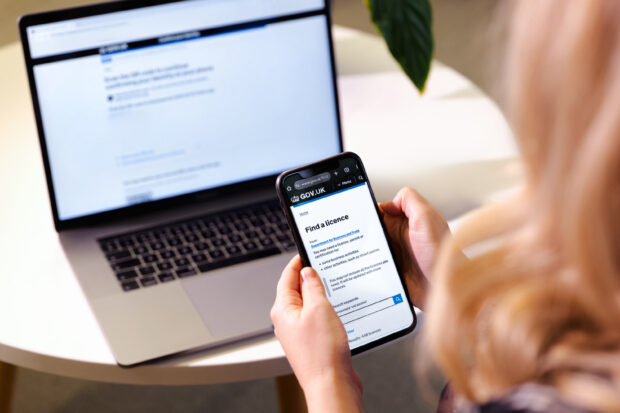 A woman holds a smartphone displaying GOV.UK’s ‘Find a licence’ page while standing in front of a laptop which is placed on a table