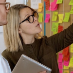 A blonde woman with glasses looking at an agile wall covered in colourful post-it notes