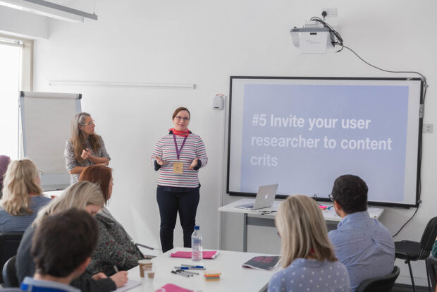 Two people standing presenting at a conference next to a screen that says, "invite your user researcher to content crits." In front of the speakers are tables of people sitting and listening and taking notes.