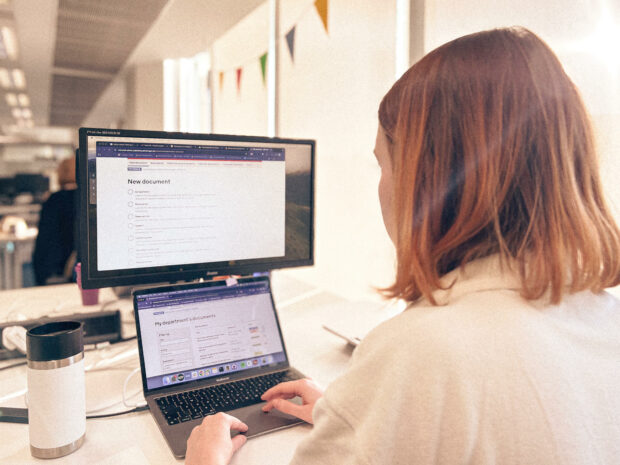 A woman sitting at a desk while using Whitehall Publisher on a laptop computer and a monitor.