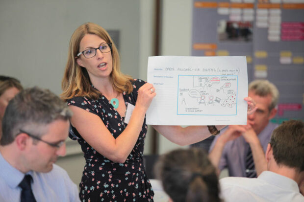 A facilitator holds up and talks through an A3 poster titled 'open policing - via digital' with a diagram of the digital process and relationships.