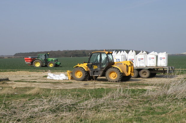 A yellow telehandler is parked beside a flatbed trailer loaded with large white bulk bags on a farm track. In the background, a green tractor with a red attachment is working a field of young crops. The landscape is flat and open with a line of trees in the distance under a clear sky.