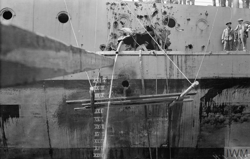The side of a QUEEN ELIZABETH class battleship in dry dock showing damage caused by a German shell at the battle of Jutland, a dock worker can be seen working inside the shell hole whilst several more are stood watching.