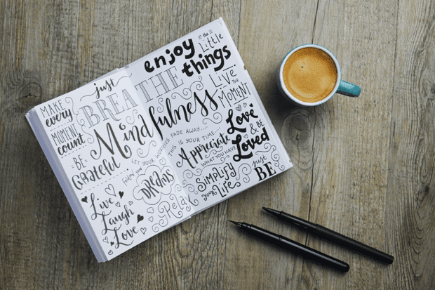 Mindfulness hand-lettered sketch notes in notepad on wooden desk with cup of coffee and pens