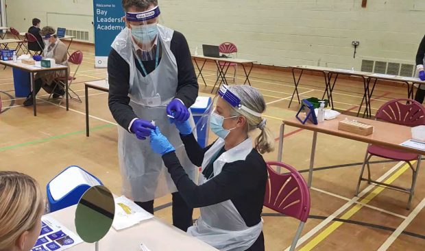 Adults wearing gloves, plastic visors and aprons sit in a sports hall, one handing a syringe to the other