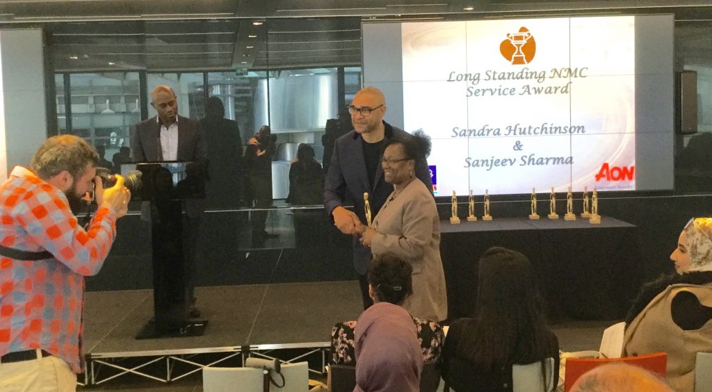 Sandra Hutchinson posing for a photograph with Paul Elliot aftre receiving her "Long Standing NMC Service Award". Her name is being projected on the screen behind her as it that of Sanjeev Sharma who also received this award.
