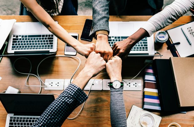 Five people sitting around a desk with laptops and folders. They are all reaching into the middle of the desk in order to fistbumping each other.