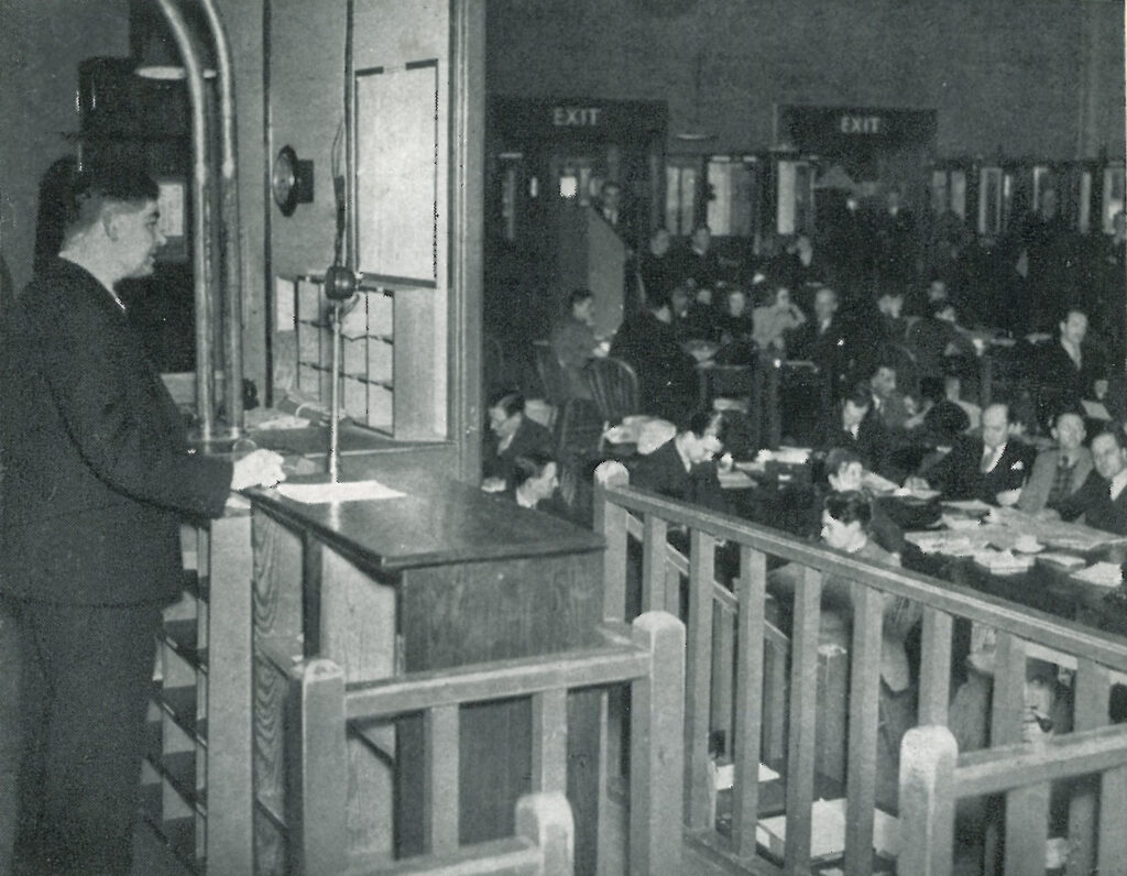 In the Ministry of Information Press Room, a man stands at a microphone, possibly making an announcement to the large number of journalists seated in the hall.