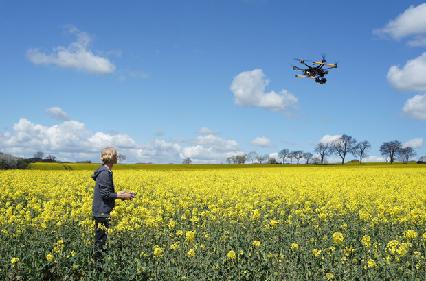 A person standing in a field of yellow flowering crops operates a handheld controller while a multi-rotor drone flies above. The field stretches to the horizon under a blue sky with scattered clouds and a line of trees in the distance.