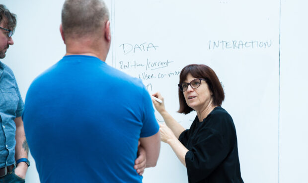 Tory standing at a white board and talking to 2 people