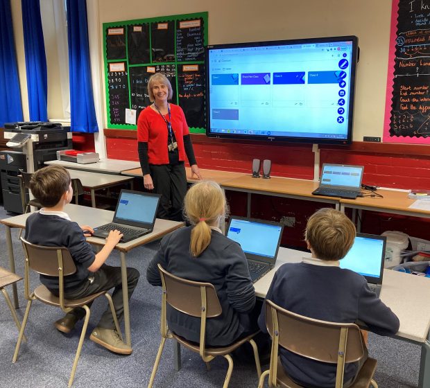A female teacher stands at the front of a classroom smiling at the camera, primary age children face h