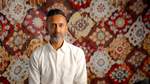 Artist Hetain Patel sits facing the camera with a colorful patterned carpet behind him