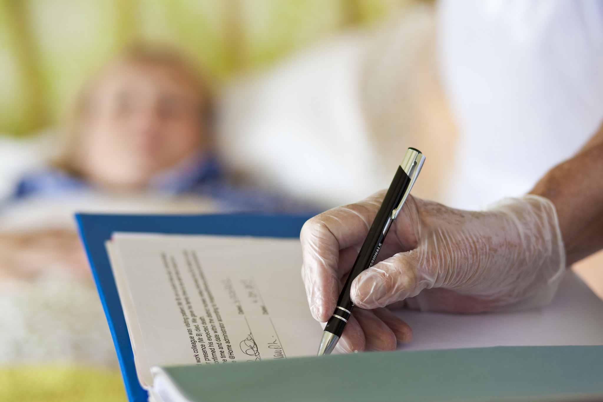 Care professional writing notes by bedside wearing surgical gloves