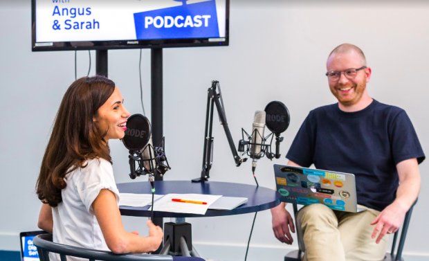 2 podcast authors sitting in the recording studio, with recording equipment on the table and a big screen behind them