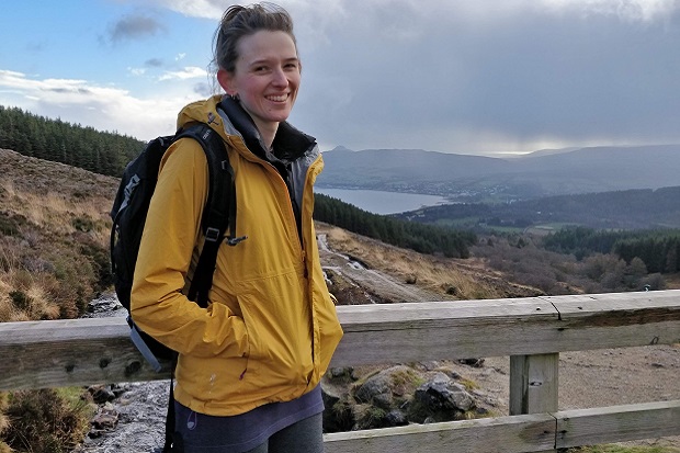 A girl wearing a yellow coat and a backpack, with hills and water in the background.