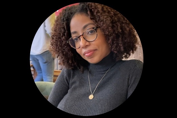 A lady with shoulder length curly hair and glasses, wearing a grey sweater and necklace.