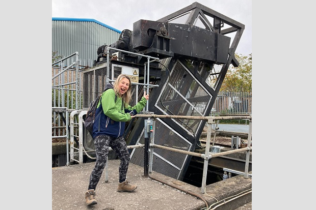 A lady, wearing a green and blue jacket and backpack, points to a large machine