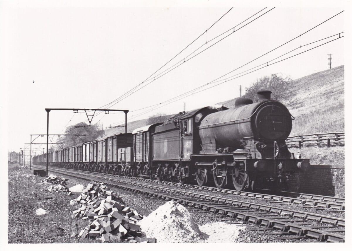 Black and white photo of steam locomotive train - E.R. Class J39 0-6-0 64827 on an up freight passing Godley East Junction May 1954