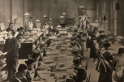 A large room full of men and women working at benches wearing white aprons