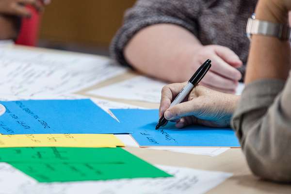 Close up of a table in a workshop with paper scattered all over. We see the arms of two people, one is making notes.