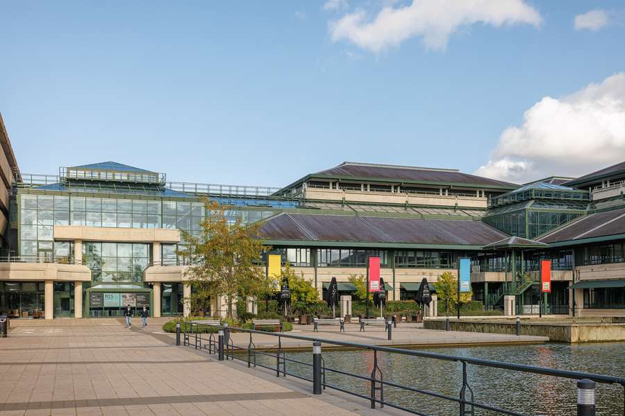A modern building with large glass windows. Colourful banners are flown outside the building on tall poles. There is also a large lake.