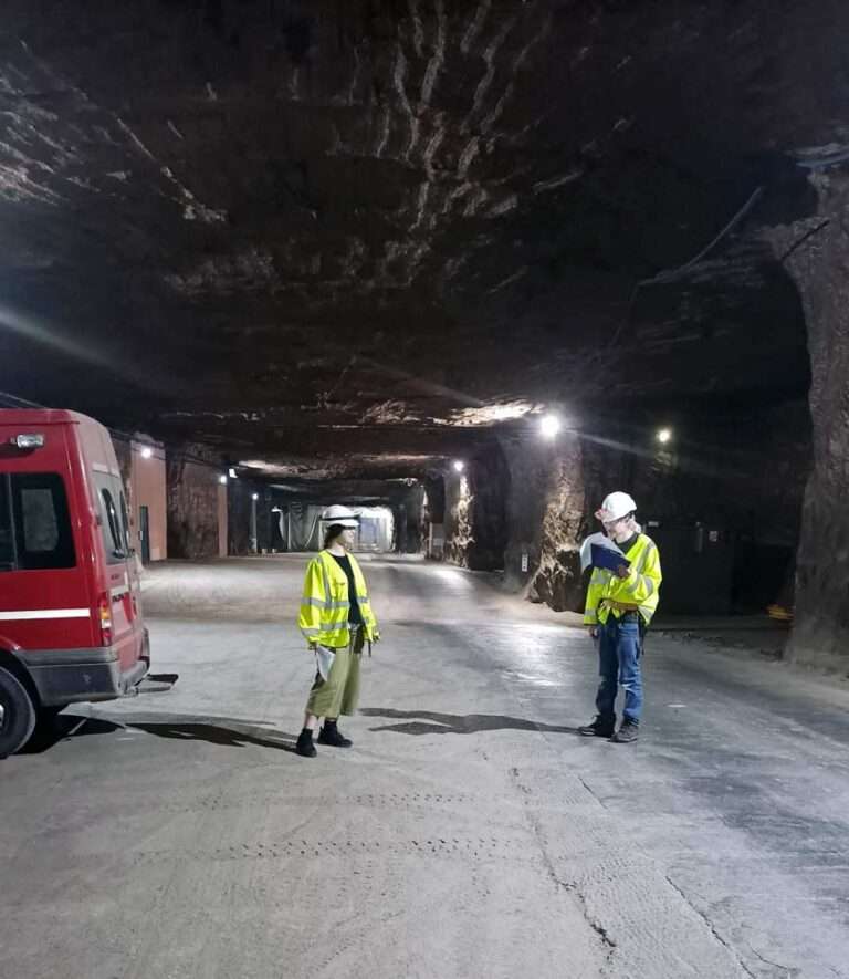 Two people in hi-vis vests and hard hats stand in a vast underground space.
