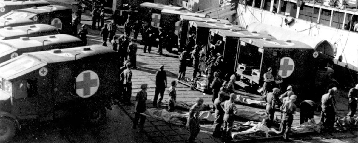 Photograph looking down on around thirty people unloading stretchers from vans marked with crosses.